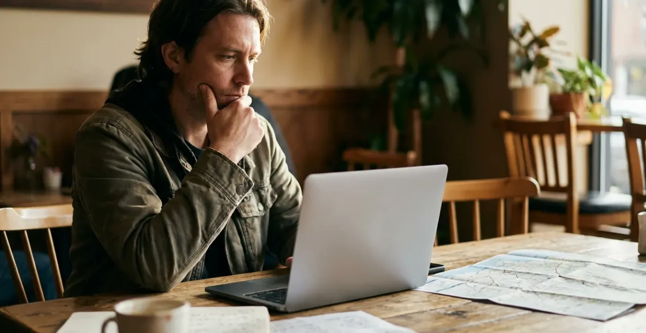 Un voyageur concentré examine des avis clients sur son ordinateur portable dans un café, avec une carte et un carnet de notes à côté
