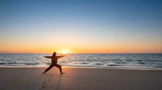 Pratique de yoga matinale face à la mer, une personne en posture du guerrier sur le sable au lever du soleil